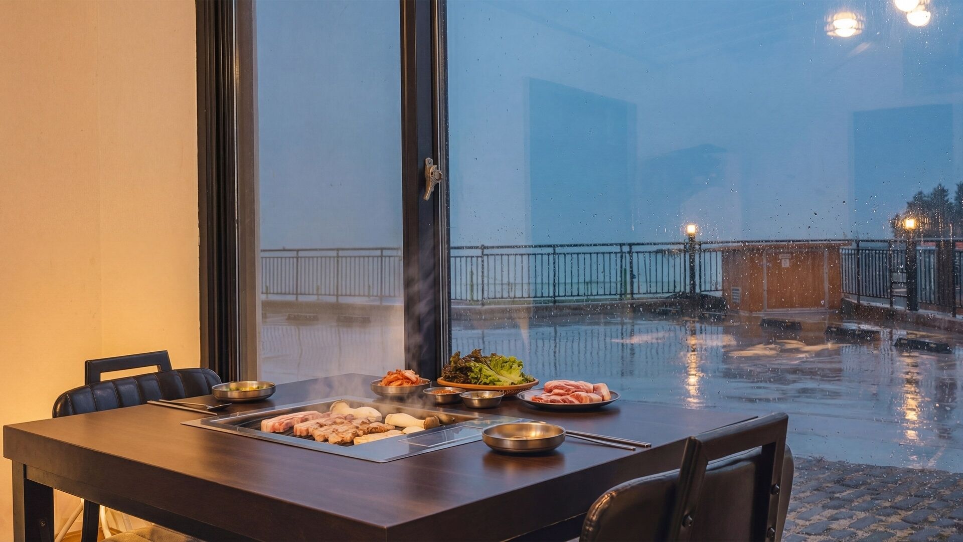 A dining table with various dishes, a view of a rainy outdoor area, and a window with a view of a building.