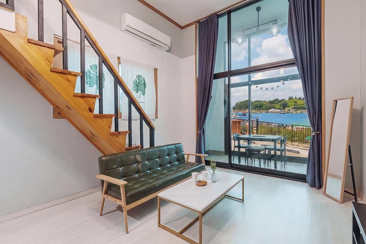Un salon moderne avec un escalier, un canapé vert, une table basse et une grande fenêtre donnant sur la mer.