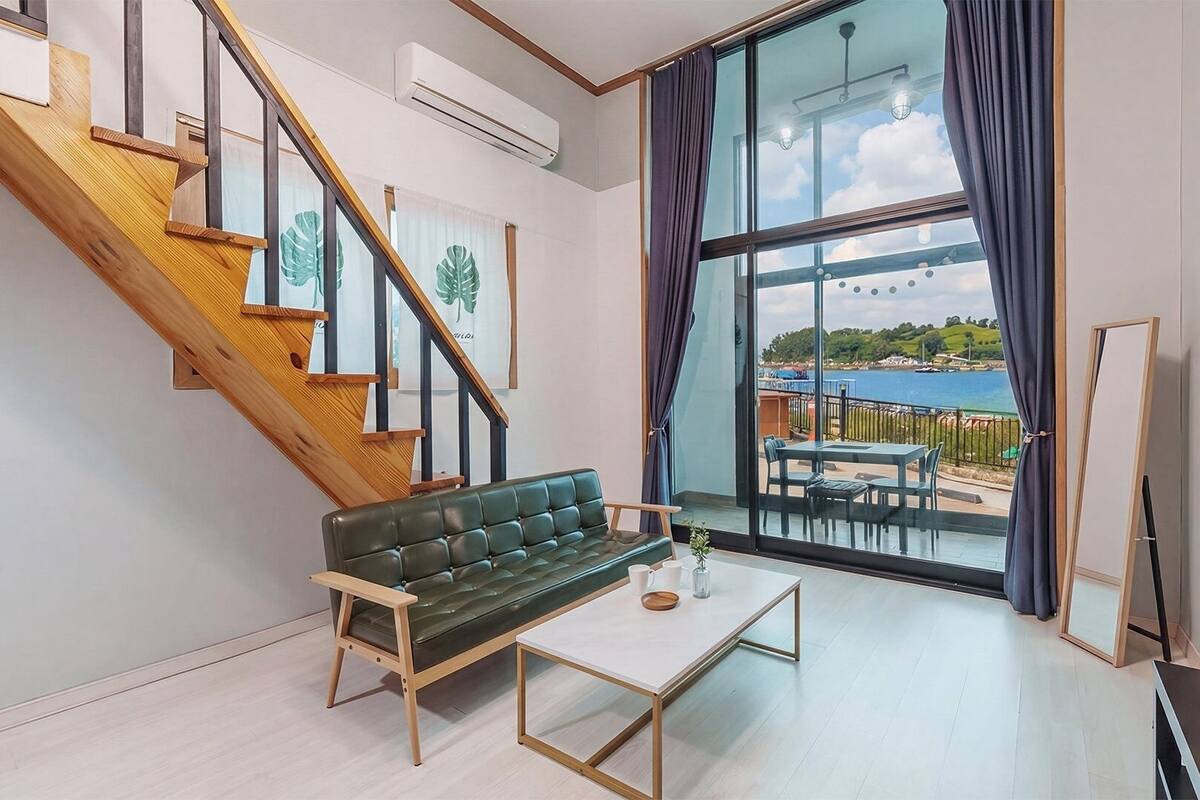 A modern living room with a staircase, a green sofa, a coffee table, and a large window with a view of the sea.