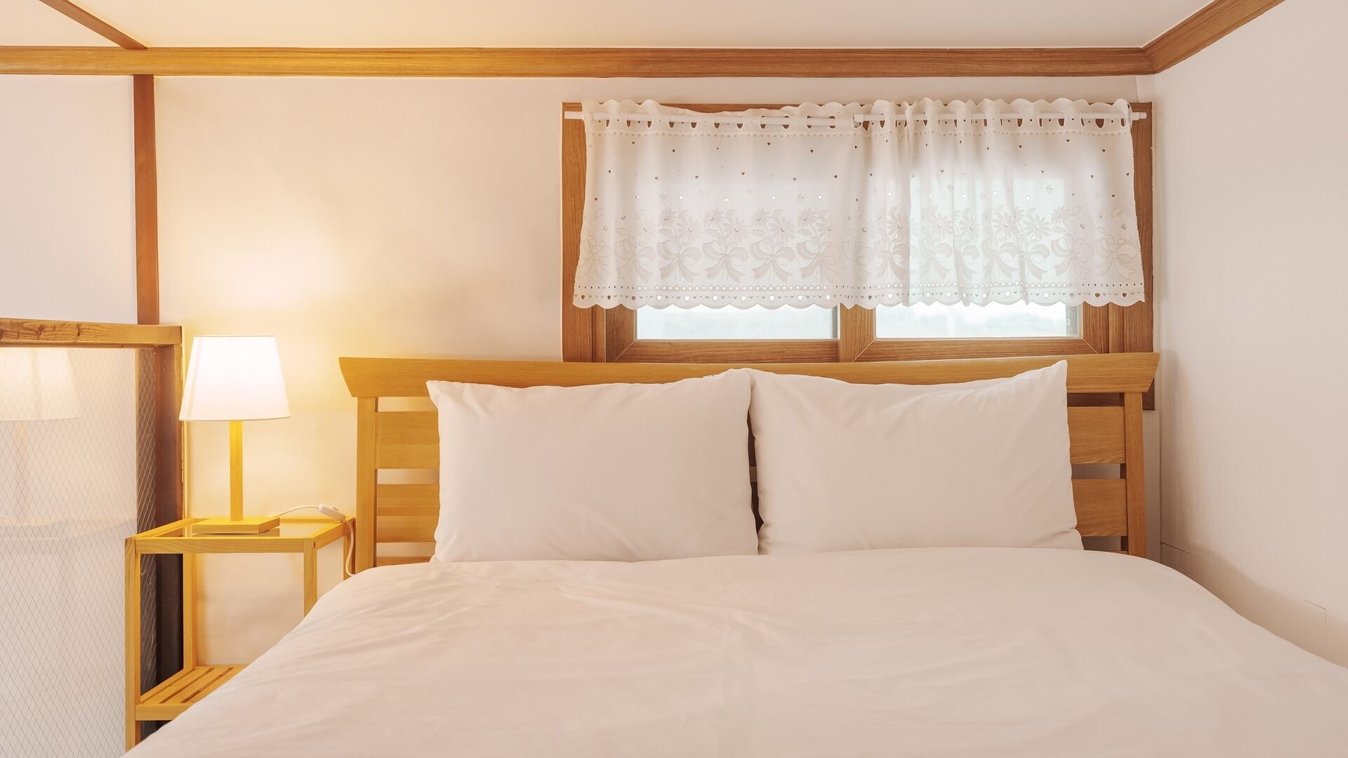 Un dormitorio con cabecera de madera, dos almohadas blancas, una lámpara de noche y una ventana con cortina de encaje.