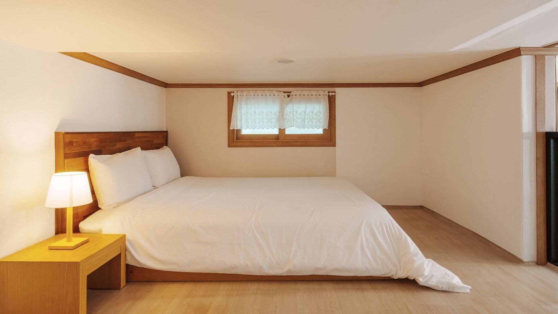 Un dormitorio con cabecera de madera, una cama individual, una mesita de noche con lámpara y una ventana con cortinas de encaje.