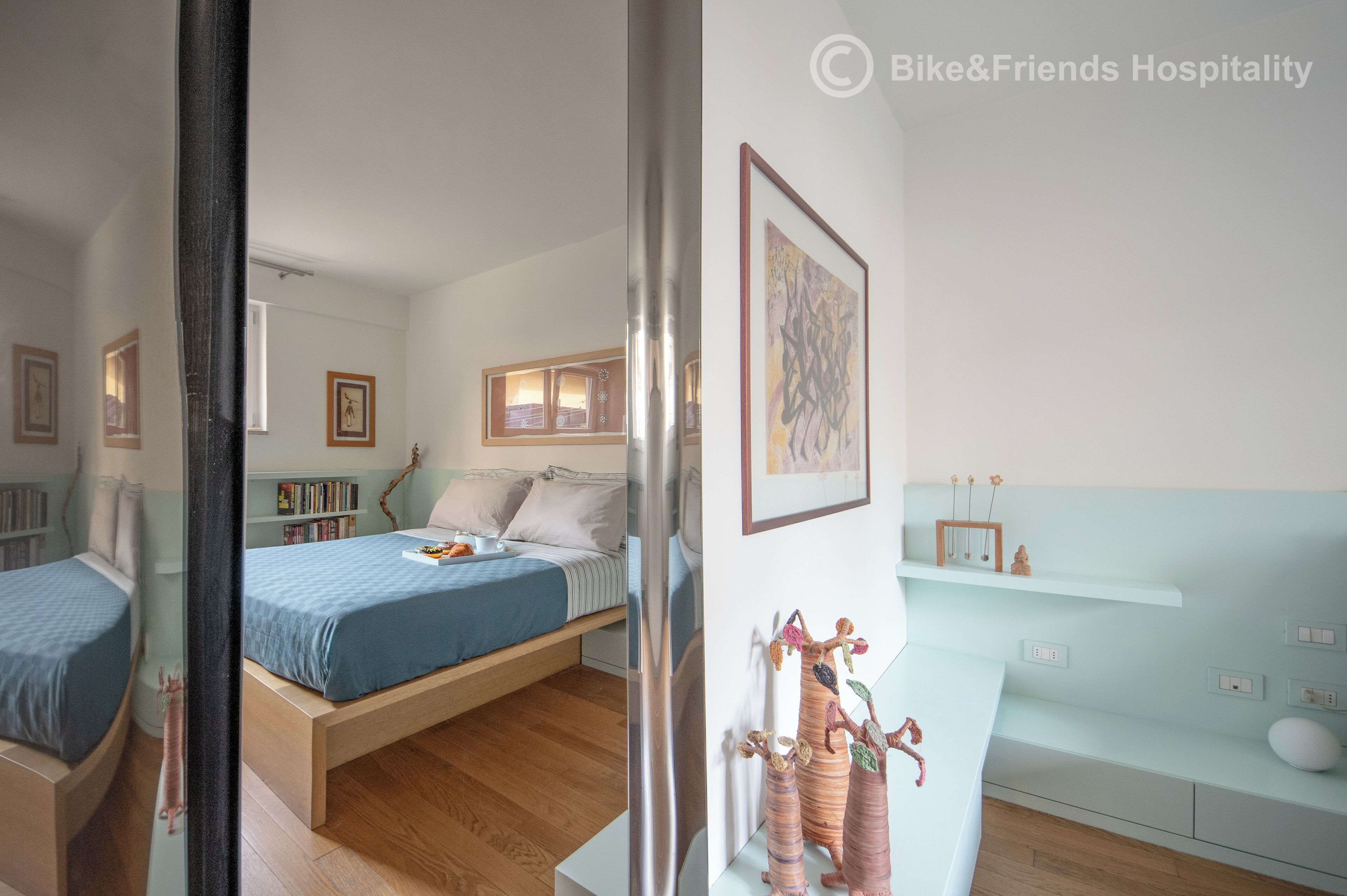 A modern bedroom with a bed, bedside tables, and a nightstand. There are framed pictures on the wall and a small shelf with decorative items.