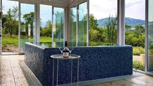 Una habitación iluminada por el sol, con un amplio ventanal, un banco de azulejos azules y una pequeña mesa con una botella y un vaso.