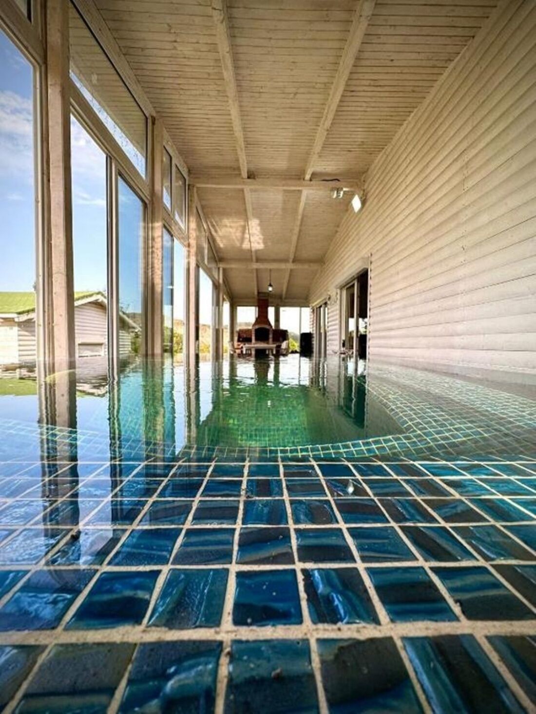 A covered swimming pool with a tiled floor, large windows, and a view of the surrounding area.