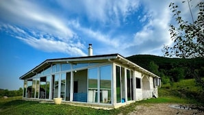 Una casa moderna con amplios ventanales y fachada de madera, rodeada por un campo cubierto de hierba y colinas.