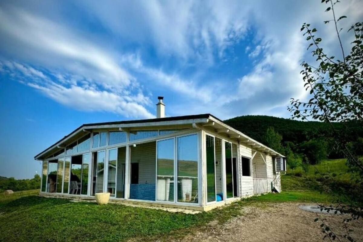 Une maison moderne dotée de grandes fenêtres en verre et d’un revêtement extérieur en bois, entourée d’un champ verdoyant et de collines.