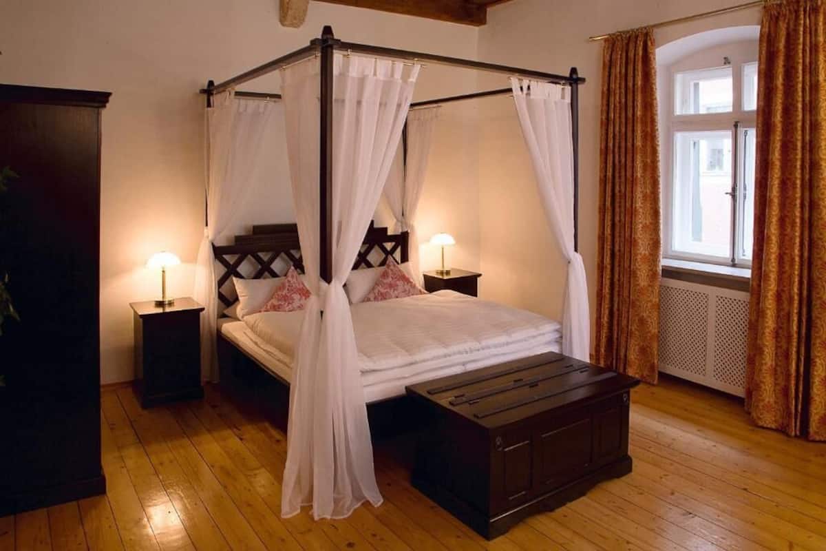 A four-poster bed with white linens and a wooden bench, two bedside tables with lamps, and a window with curtains.