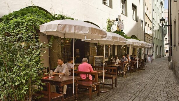 Outdoor seating with umbrellas, a cobblestone street, and buildings with signage. - Dicker Mann Hotel Zum Blauen Krebs (Regensburg)