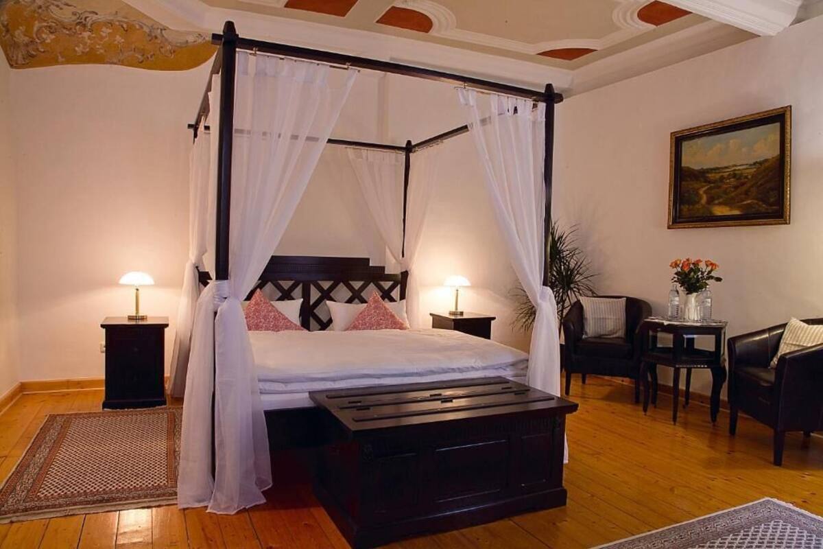 A four-poster canopy bed with white linens, two bedside tables with lamps, a dark wooden bench, a small table with flowers, and a painting on the wall.