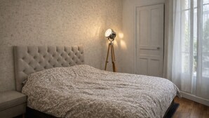 Slaapkamer met een gestoffeerd hoofdeinde, een bed met een decoratieve spreidek, een staande lamp, een houten vloer en een deur.