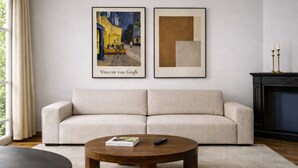 Een woonkamer met een beige bank, een ronde houten salontafel en twee ingelijste kunstwerken aan de muur.