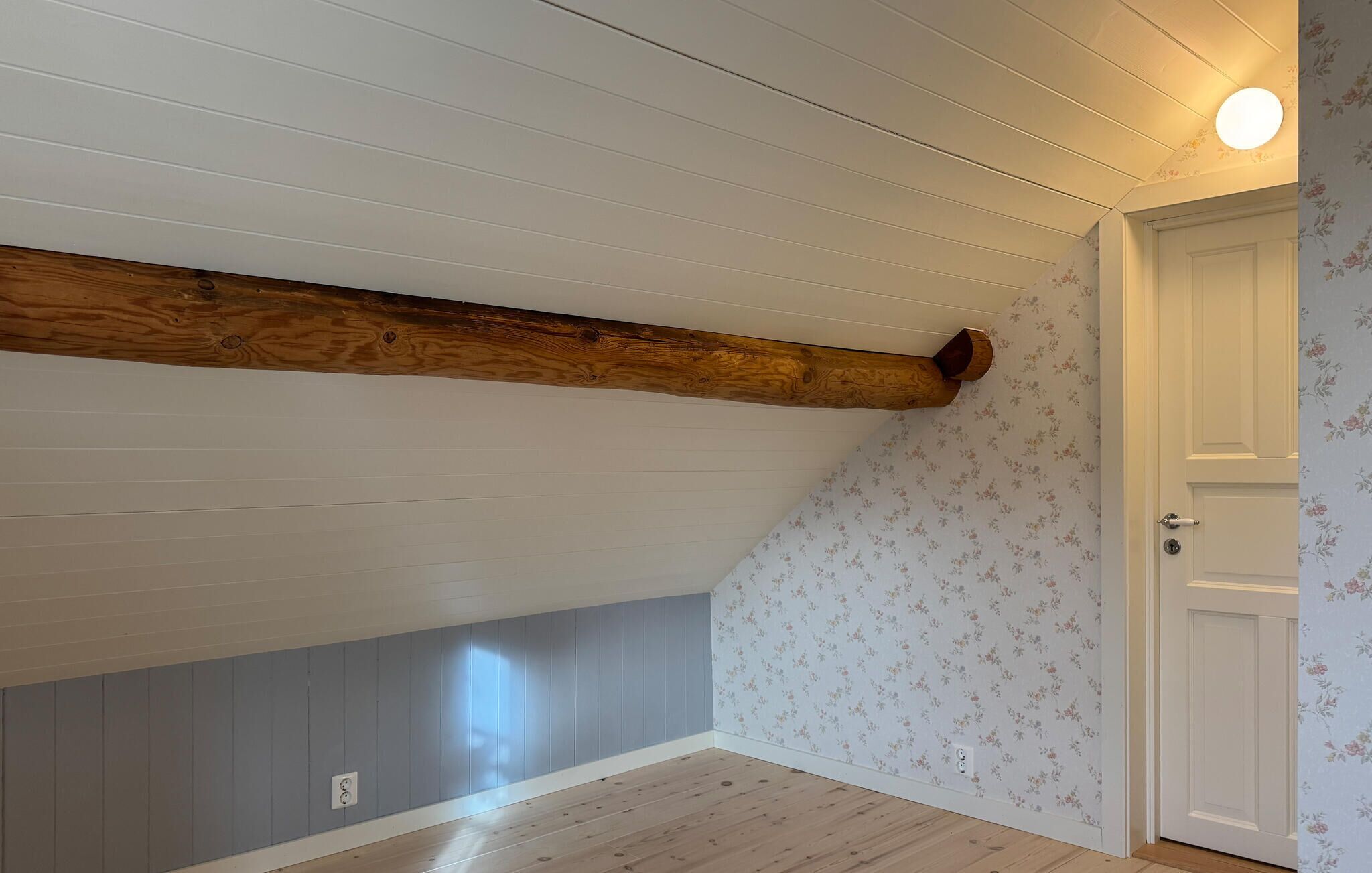 A room with a sloped ceiling, wooden beam, floral wallpaper, and a door.