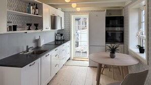 A modern kitchen with white cabinets, a black countertop, and a built-in oven. There is a dining area with a round table and chairs, and a potted plant on the windowsill. - Beach front home in Vikebygd with WiFi (Vikebygd)
