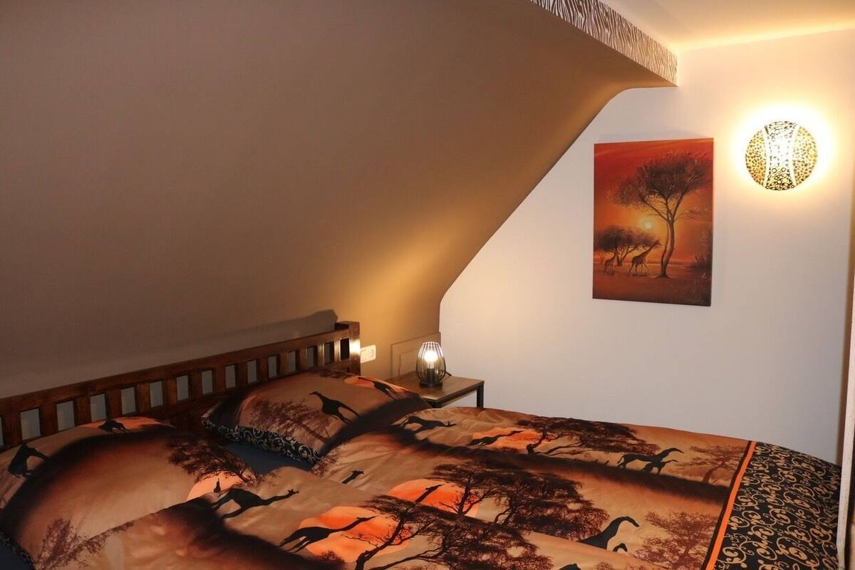 Een slaapkamer met een schuine kap, een bed met een geborduurde spreideken, een wandlamp en een schilderij van een zonsondergang.