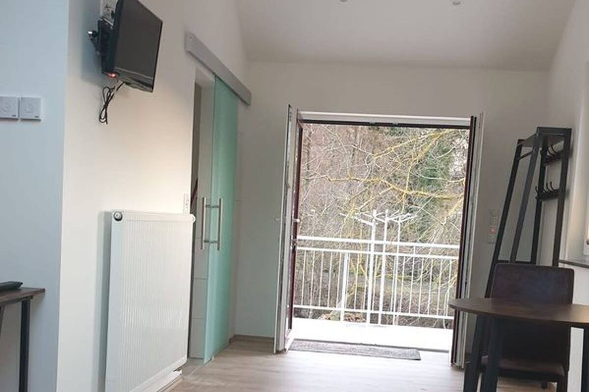 Een kamer met een radiator, een aan de muur bevestigde televisie, een eettafel met stoelen en een balkon met een metalen hekwerk.