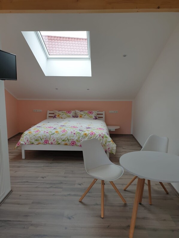 A bedroom with a skylight, a bed with floral bedding, a white chair, and a small table. - Schlafwelt Schalkhaus (Elsenfeld)