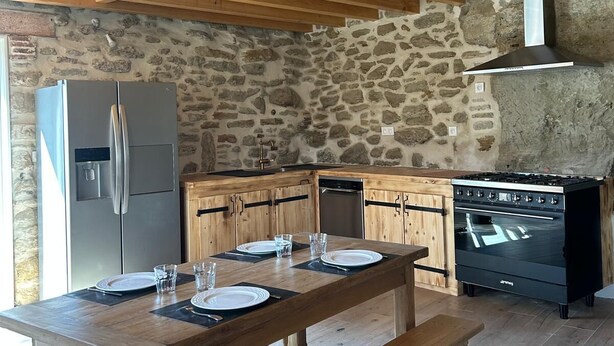 A rustic kitchen with stone walls, wooden beams, a stainless steel refrigerator, a stove, and a dining table set for four. - NATURE & SERENITY GITE (Saint-Avit)
