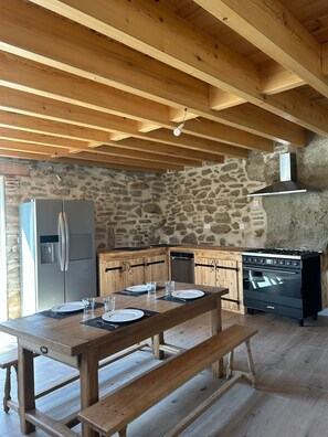 A rustic kitchen with stone walls, wooden beams, a stainless steel refrigerator, a stove, and a dining table set for four. - NATURE & SERENITY GITE (Saint-Avit)
