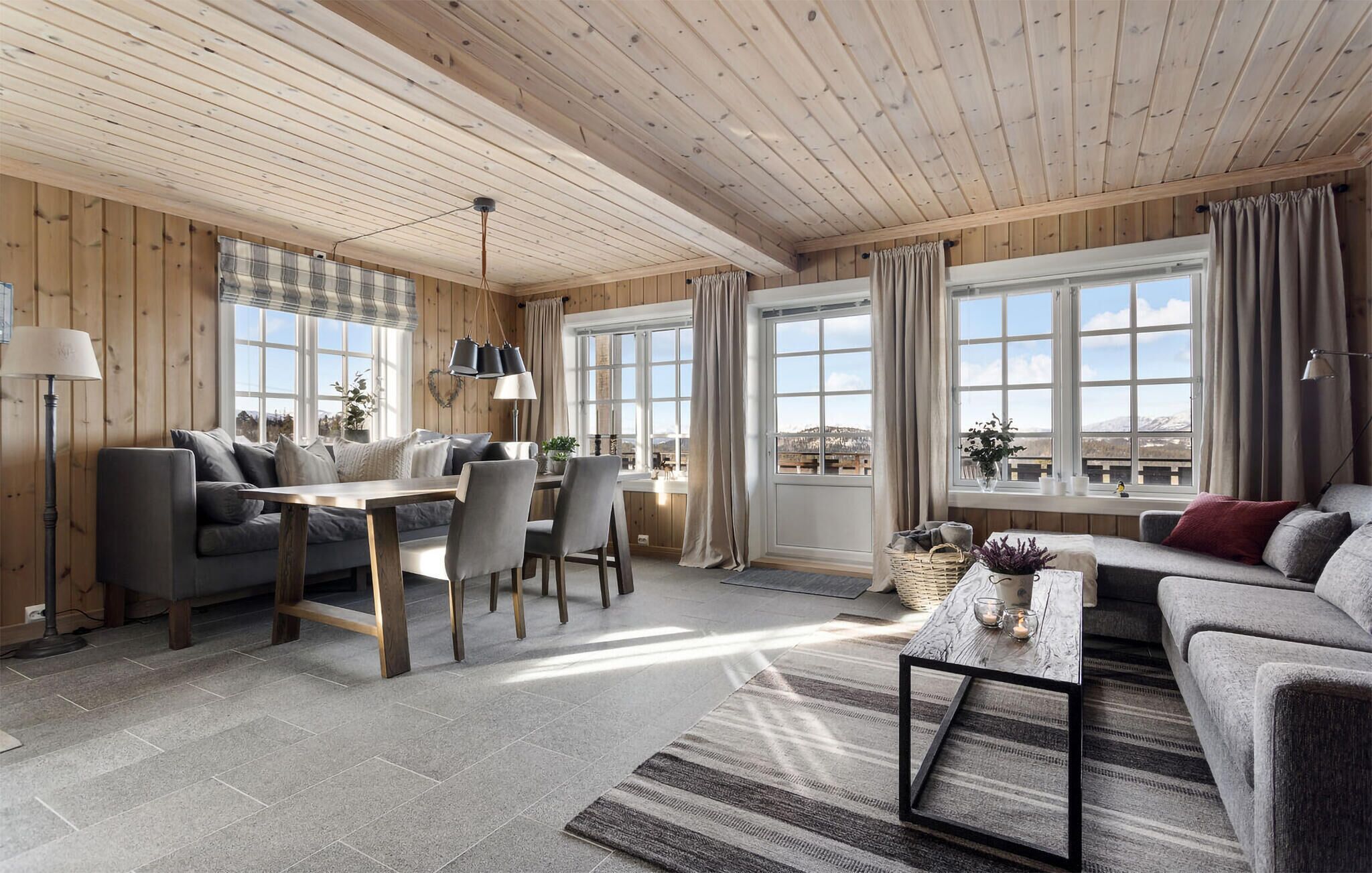 A spacious living area with a wooden ceiling, large windows, a dining table, and a sofa.