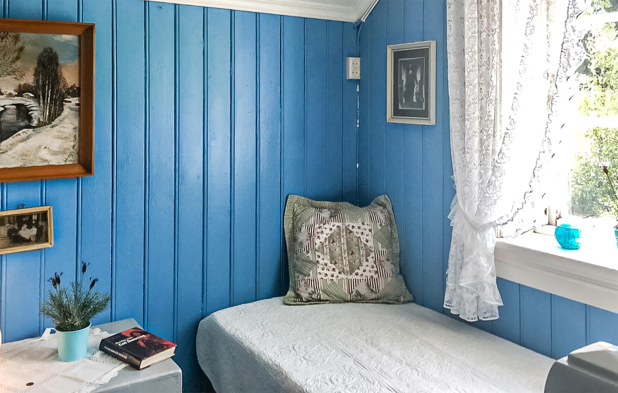 A room with blue walls, a bed with a white bedspread, a pillow with a floral pattern, a small table with a plant and a book, and a window with a white curtain.