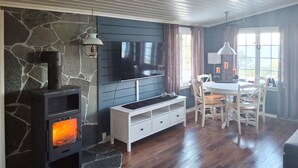 Una sala de estar acogedora con una chimenea de piedra, un televisor de pantalla plana, una consola blanca y un comedor con sillas de madera.