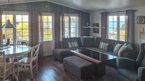 Una sala de estar acogedora con un sofá, un reposapiés y una mesa de centro de vidrio. Hay una zona de comedor con una mesa y sillas. Amplias ventanas ofrecen vistas al exterior.