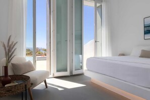 Ein modernes Schlafzimmer mit einem großen Bett, einer Sitzecke inklusive Sessel und kleinem Tisch sowie Ausblick ins Freie durch Glastüren.