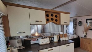 Une cuisine compacte équipée d’un grille-pain, d’une bouilloire et d’une machine à café sur le plan de travail. Il y a une fenêtre avec des stores et une horloge au mur.