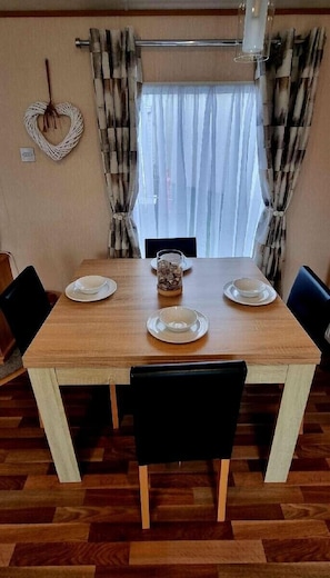 Una mesa de comedor con cuatro sillas, un jarrón con flores secas y una ventana con cortinas traslúcidas.