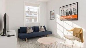 Un salon moderne avec un canapé bleu, une table basse ronde et un téléviseur posé sur un meuble. Un poster orne le mur et une fenêtre laisse entrer la lumière naturelle.