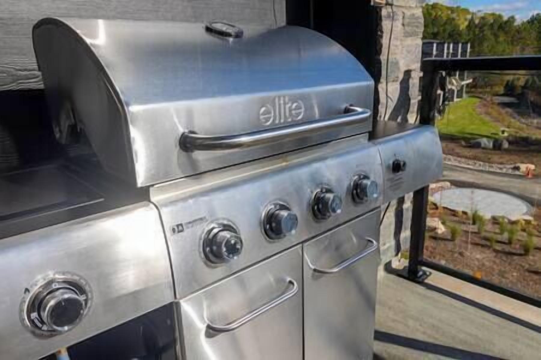A stainless steel grill with four burners and a lid, located outdoors with a view of a landscaped area.