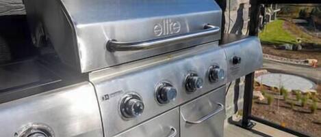 A stainless steel grill with four burners and a lid, located outdoors with a view of a landscaped area.