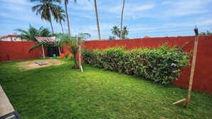 Un jardin bien entretenu, avec un mur rouge, de l’herbe verte et une variété de plantes, dont un arbuste aux fleurs violettes.