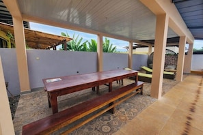 An outdoor patio area with a wooden bench and a table, surrounded by a low wall and a covered roof structure.