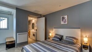 A modern bedroom with a large bed, bedside lamps, a small table, and a view into the kitchen area. - "Villa Athena" Cottage with Shared Pool, Private Terrace, and Wi-Fi (Montagnac)
