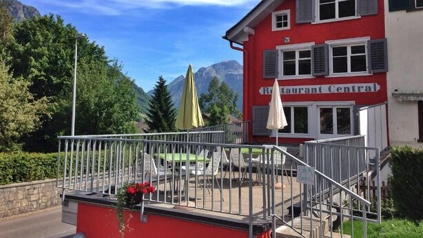 A red building with a sign reading "Restaurant Central" and outdoor seating with umbrellas. - Hotel Central (Glarus Sued)