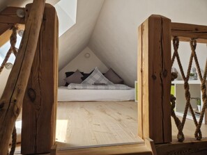 Une chambre à coucher aménagée dans un grenier, avec un plafond en pente, un lit recouvert d’une couette à carreaux et une échelle en corde.