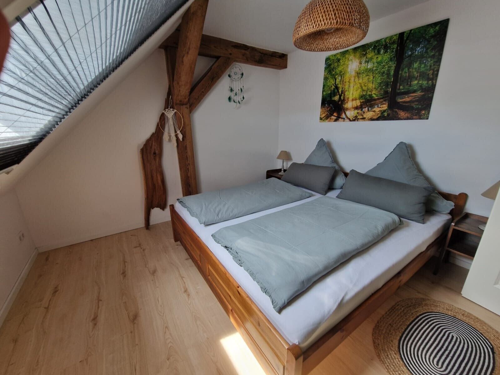 A bedroom with a sloped ceiling, a bed with grey bedding, a woven light fixture, a dreamcatcher, and a forest-themed painting.