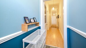 Un couloir aux murs bleus, avec un plancher en bois et un banc blanc. Des tableaux encadrés sont disposés sur une étagère, et une porte se trouve au fond du couloir.