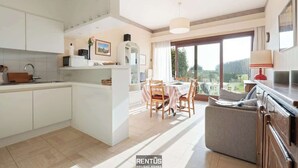 Eine moderne Küche mit weißen Schränken, ein Essbereich mit Tisch und Stühlen und Blick ins Grüne durch große Fenster.