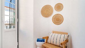 Een kamer met een witte deur, een raam met uitzicht op een brug, een houten stoel met een gevlochten zitting, en drie wanddecoraties van geweven materiaal.