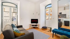 Een moderne woonkamer met een grijze bank, een blauwe hockerkruk, een Netflix-televisie en een keuken die door een open deur zichtbaar is.