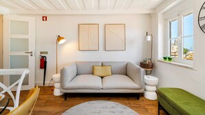 A living room with a grey sofa, a green ottoman, a round rug, a white door, and a window with a view. - Alfama Retreat Steps from Santa Luzia Viewpoint (Lisbon)