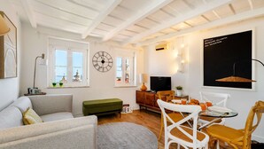 A living room with a sofa, a round rug, a dining table with chairs, a television, and a clock on the wall. - Alfama Retreat Steps from Santa Luzia Viewpoint (Lisbon)