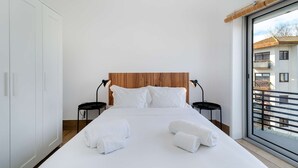 Un dormitori modern amb un capçal de llit de fusta, roba de llit blanca, dues taules de nit amb làmpades i vista a un edifici a través d'una finestra gran.