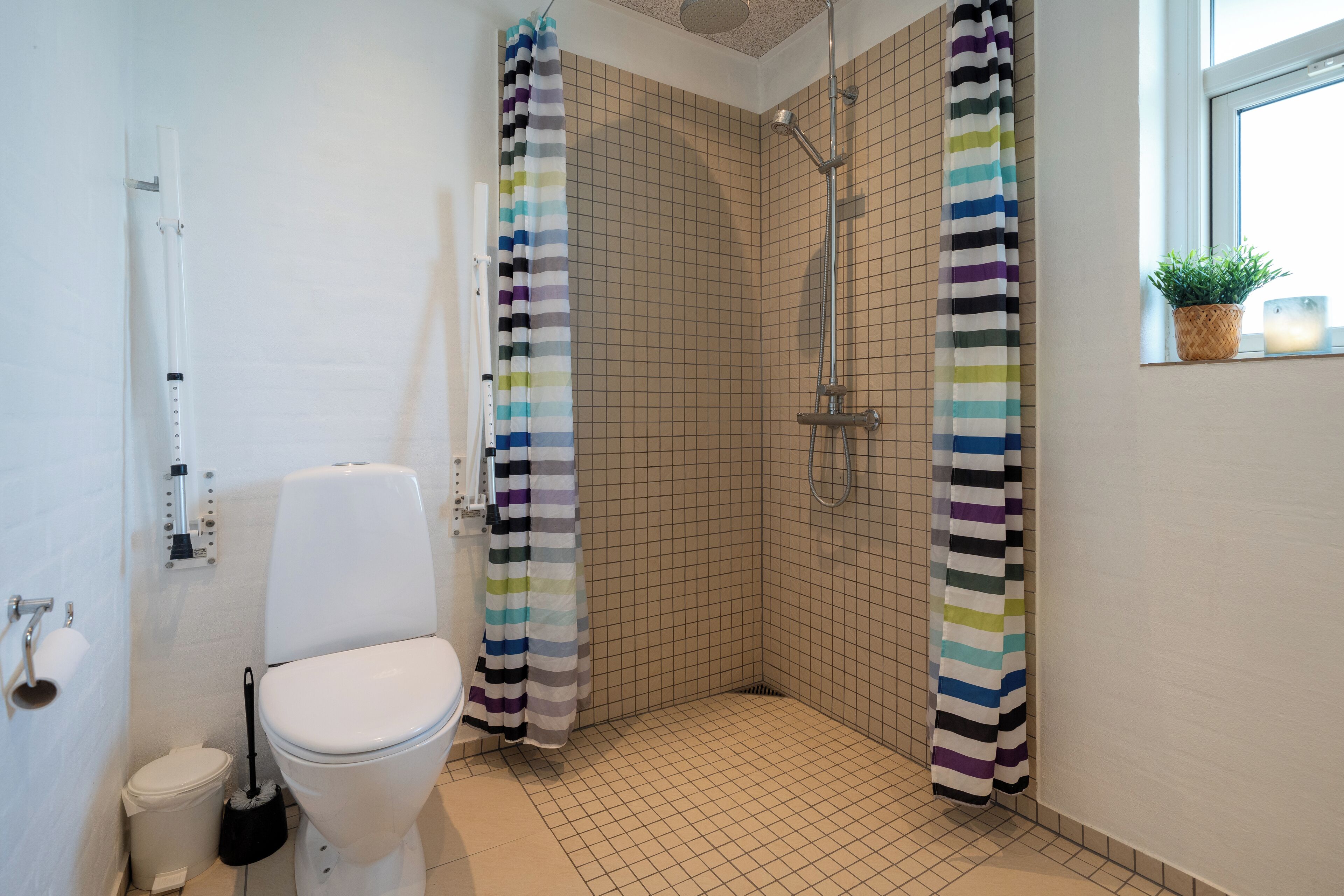 A bathroom with a toilet, a shower with a striped curtain, and a potted plant by the window.