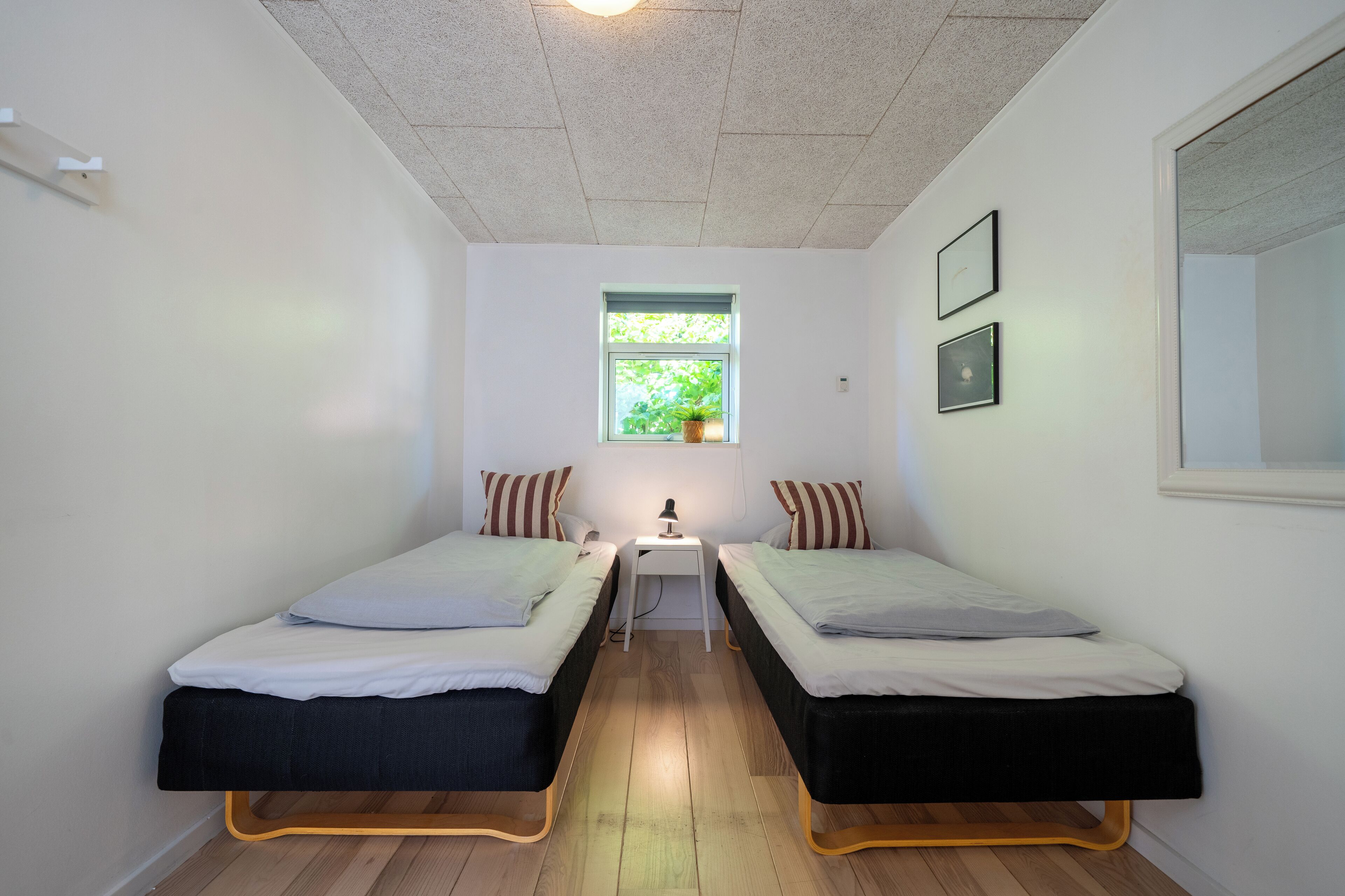 A small, well-lit room with two single beds, a small table with a lamp, a window with a plant, and framed pictures on the wall.
