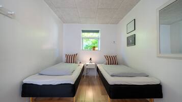 A small, well-lit room with two single beds, a small table with a lamp, a window with a plant, and framed pictures on the wall.