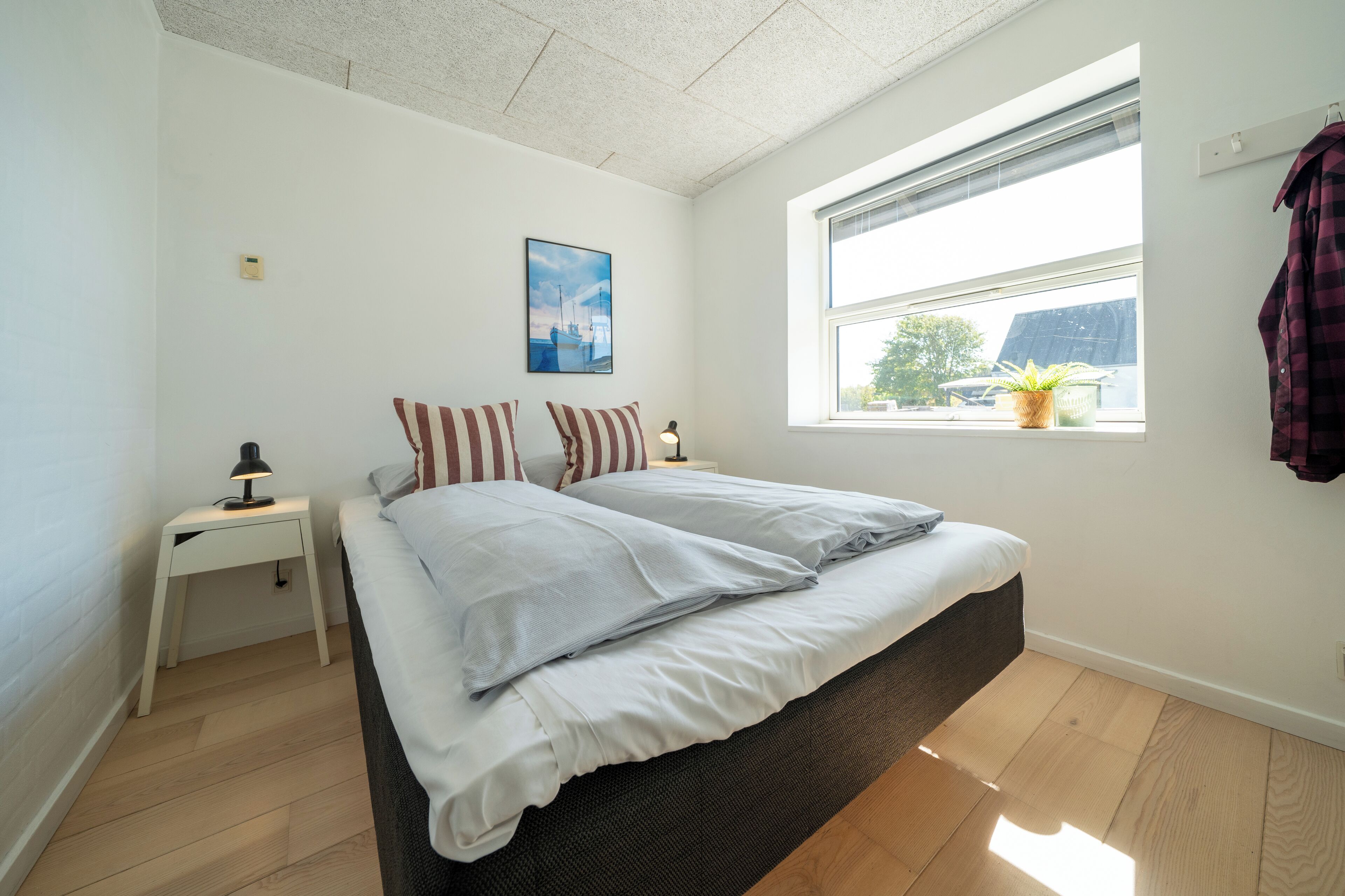 A bedroom with a bed, a nightstand, a lamp, a window with a view, and a painting on the wall.