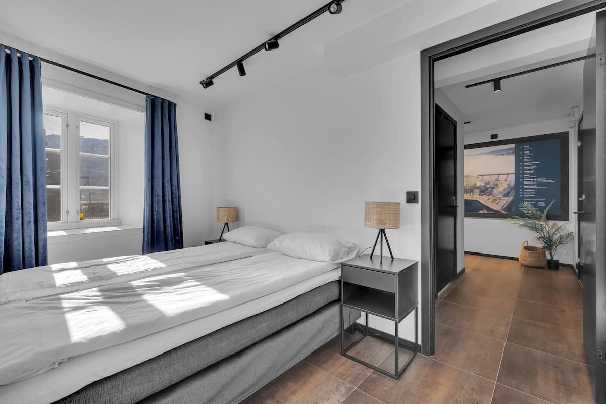 Un dormitorio moderno con cama, lámparas de noche y vistas a un paisaje urbano a través de un ventanal abierto.